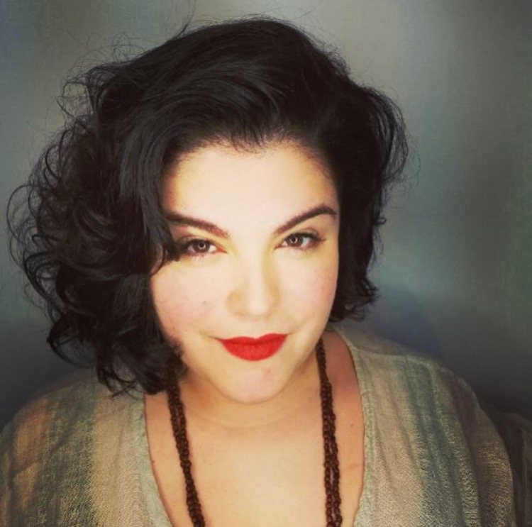 A woman with curly black hair and bold red lipstick gazes confidently at the camera, wearing a neutral-toned outfit and a bead necklace.