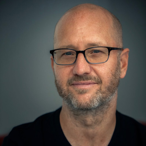 A man with a bald head and beard, wearing glasses, smiles softly against a blurred background.