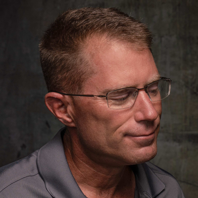 A man with short hair and glasses, wearing a gray shirt, looks thoughtfully to the side against a textured backdrop.